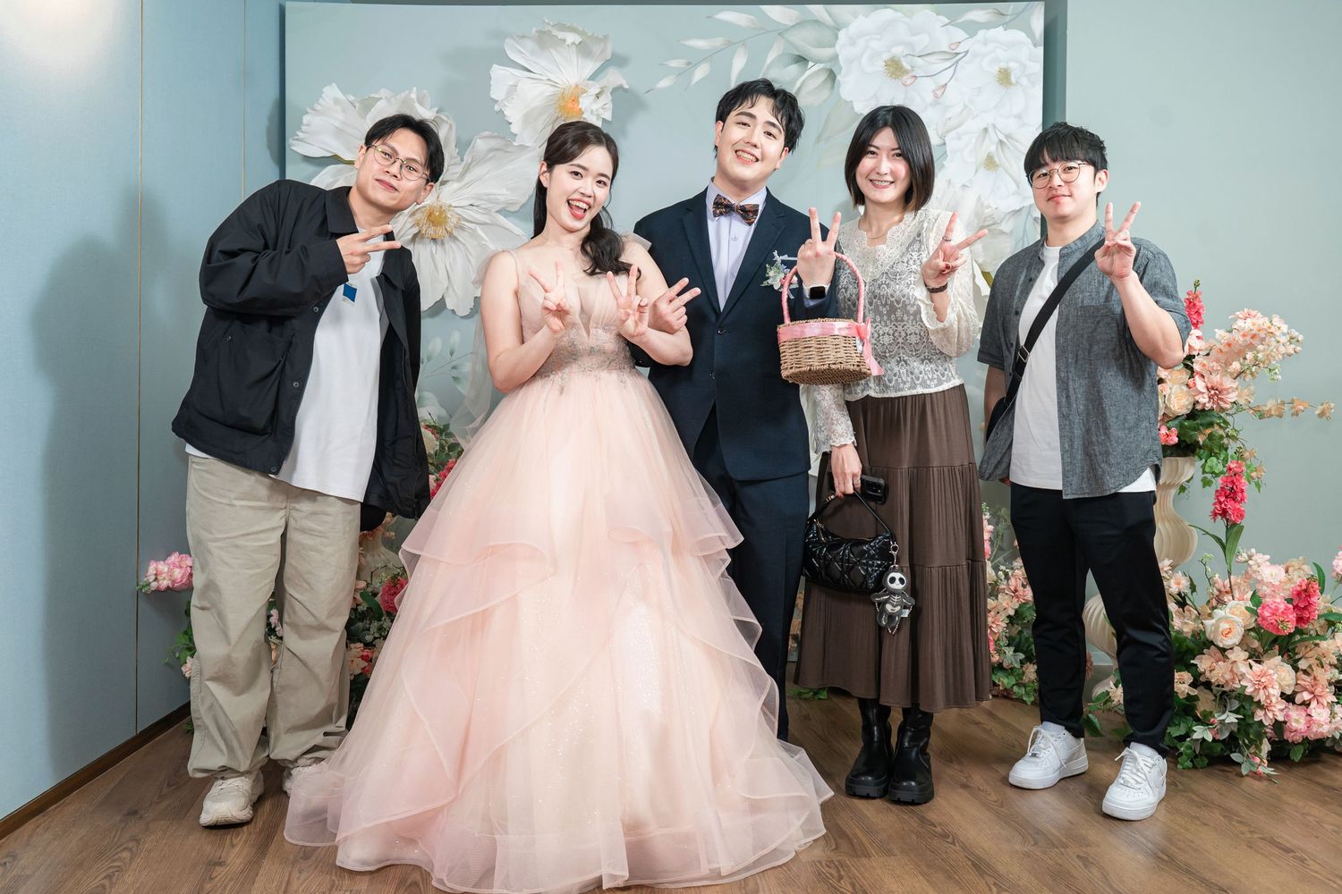 A group of people pose together in formal attire against a light blue wall decorated with pink floral arrangements.