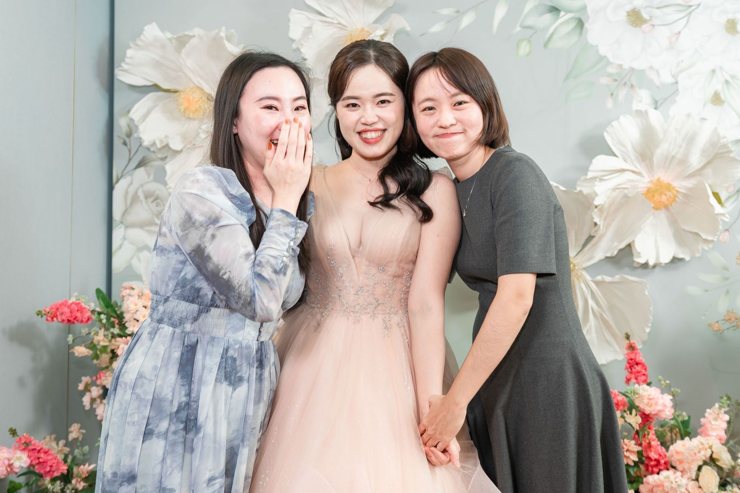 Three friends share a joyful moment together against a floral backdrop with white lilies and pink accents.