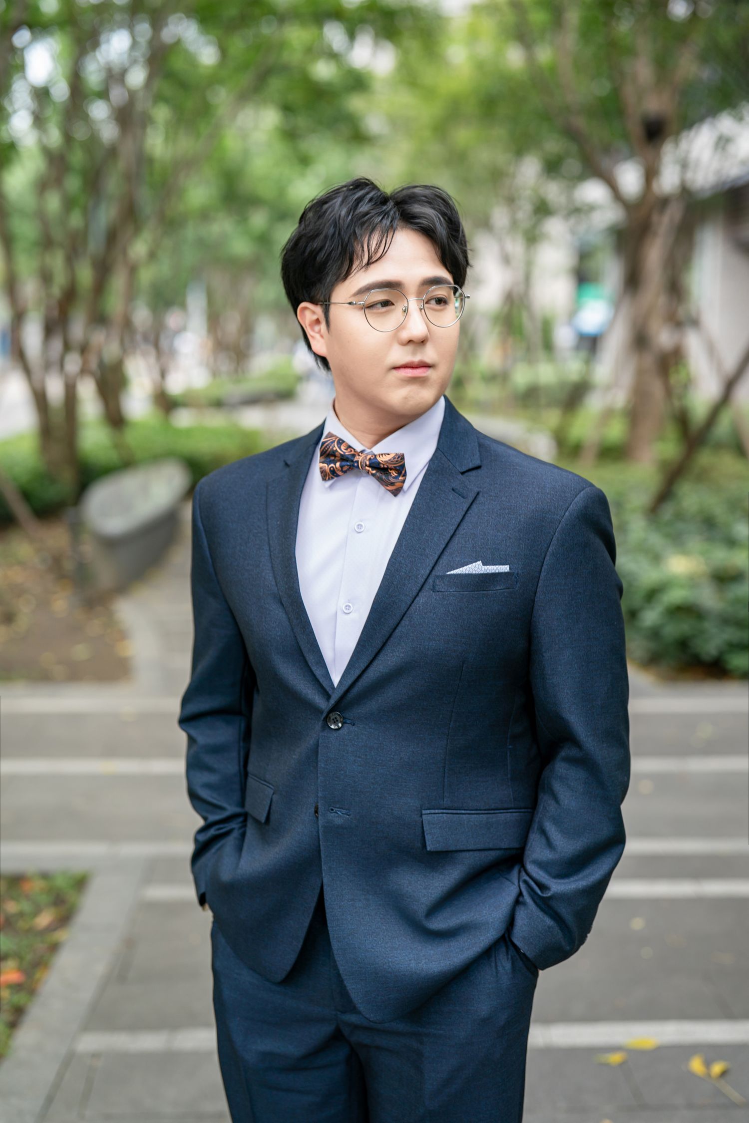 A person wearing a navy suit and bowtie poses outdoors in a garden setting for formal portraits.