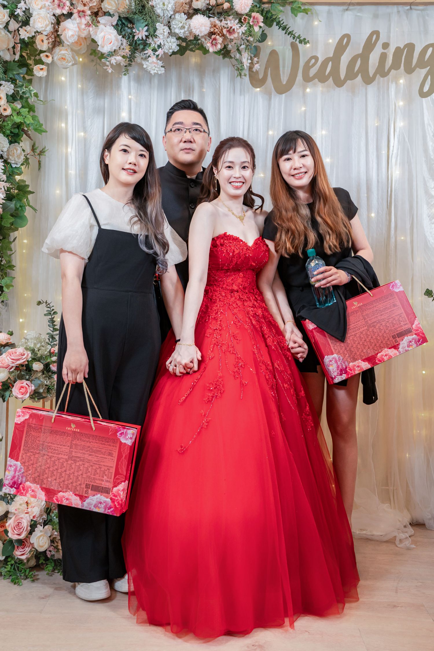Group poses at wedding celebration with decorative backdrop and red formal attire holding red gift bags.