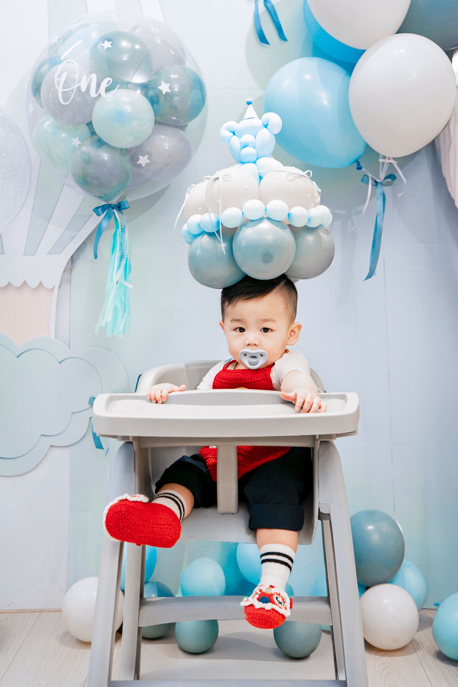 Baby's first birthday party setup with blue and white balloons and high chair against cloudy backdrop decor.