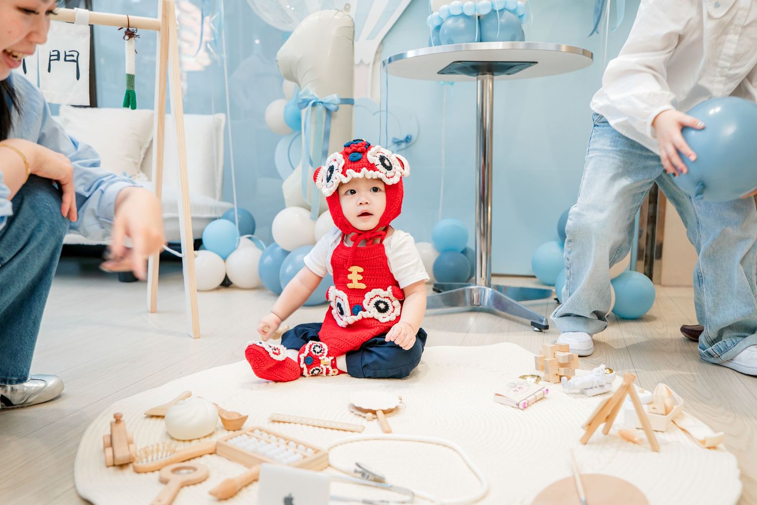 A small figure in a red and white costume sits on the floor surrounded by wooden blocks and party decorations.