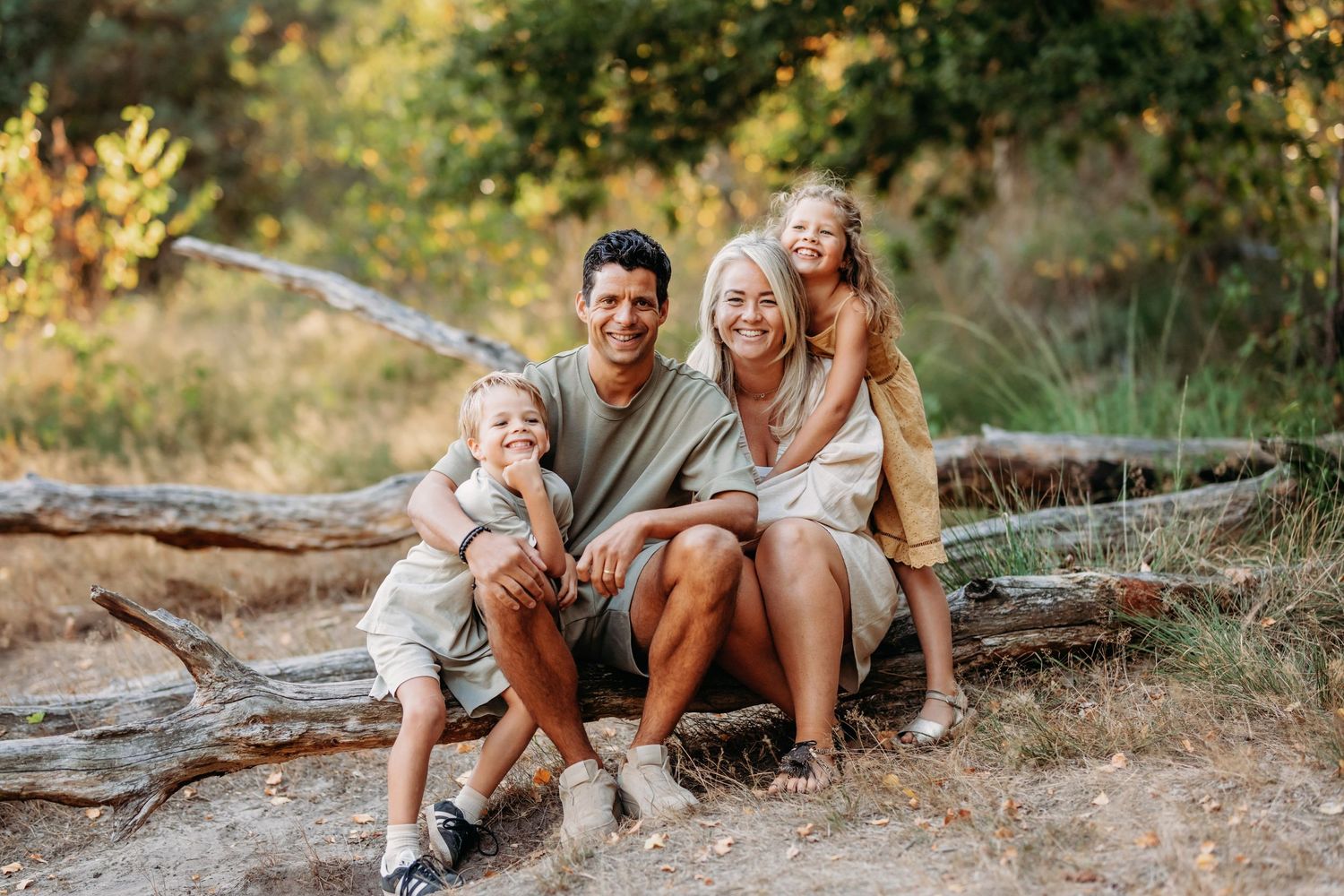 Family of four smiling and sitting on a fallen log outdoors in a sunny forest setting.