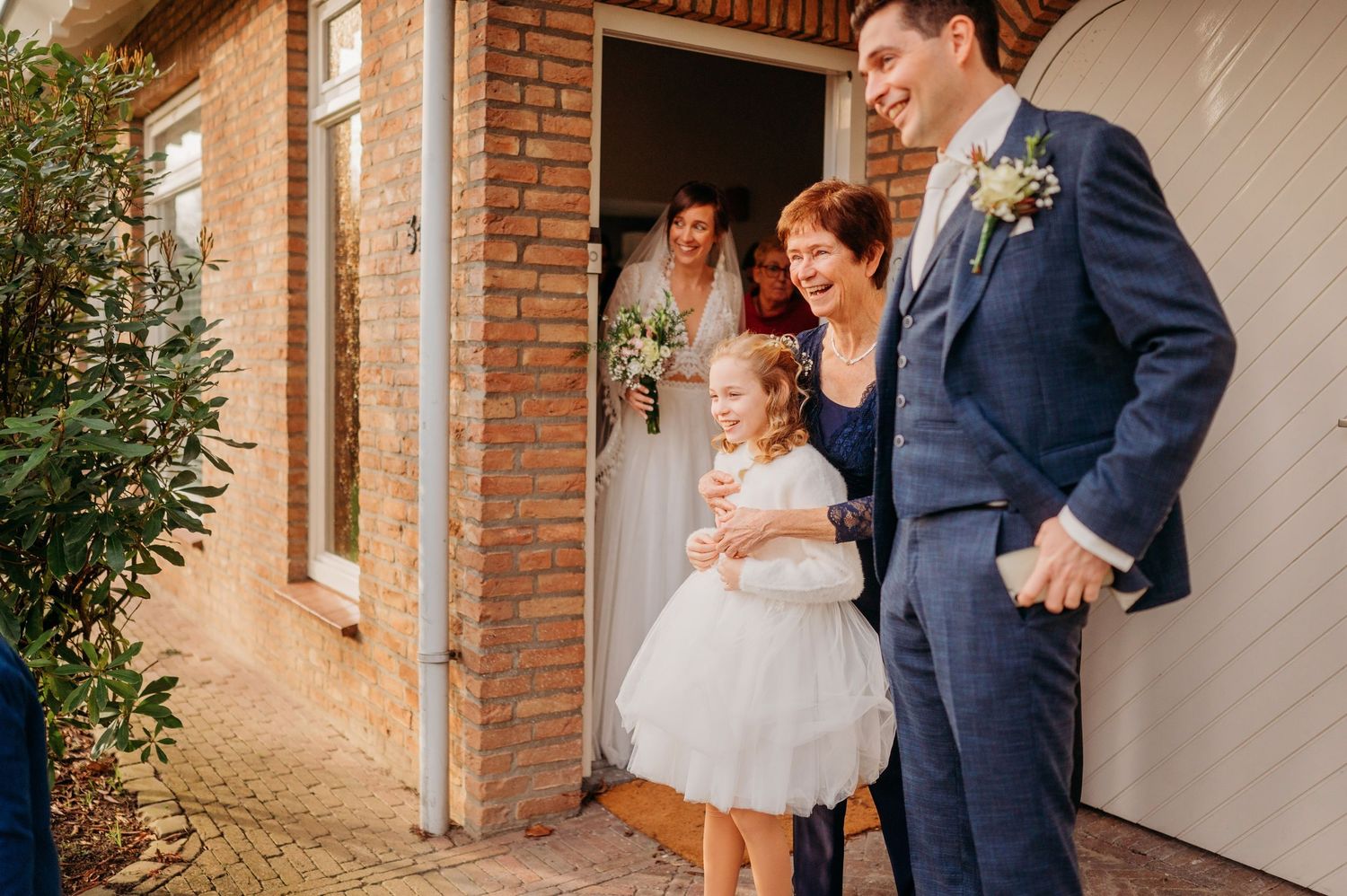 Bruidspaar en gasten delen emotioneel moment bij entree van bakstenen pand, bruidsfotografie bij Boscafé Oisterwijk, Brabant.