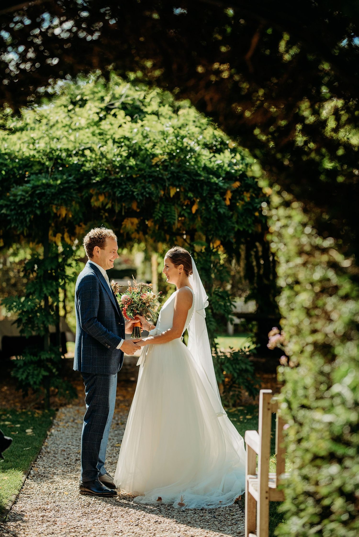 Bruidspaar onder weelderige tuinboog op grindpad tijdens gouden uur, bruidsfotografie Brabant