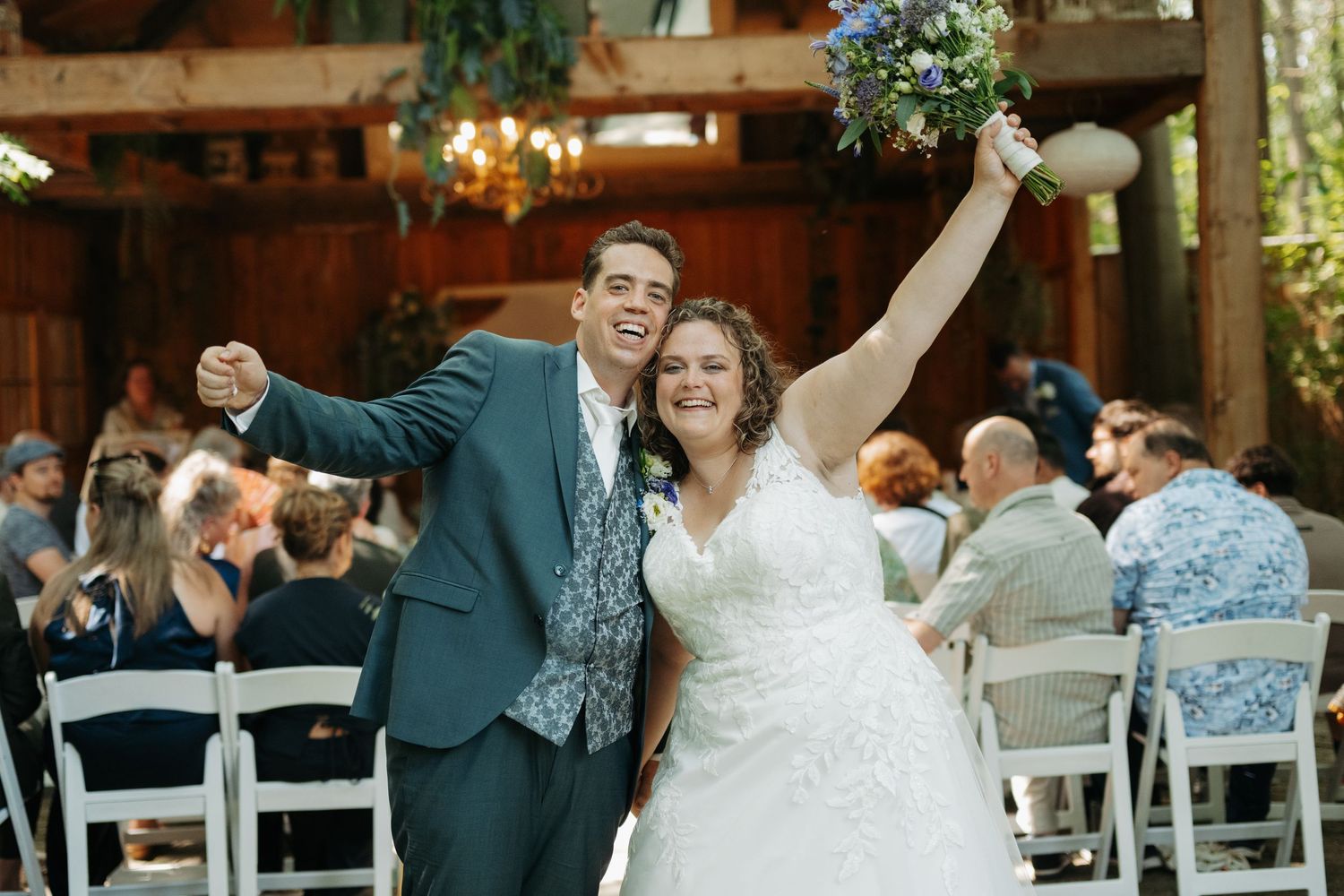 Joyful newlyweds celebrate after outdoor wedding ceremony, bride in white gown raising bouquet, groom in teal suit.