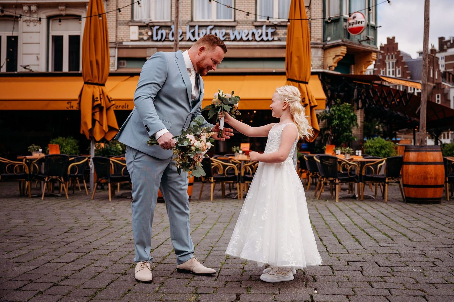 Groom in blue suit holds bride's bouquet on cobblestone square with European cafe terrace in background.