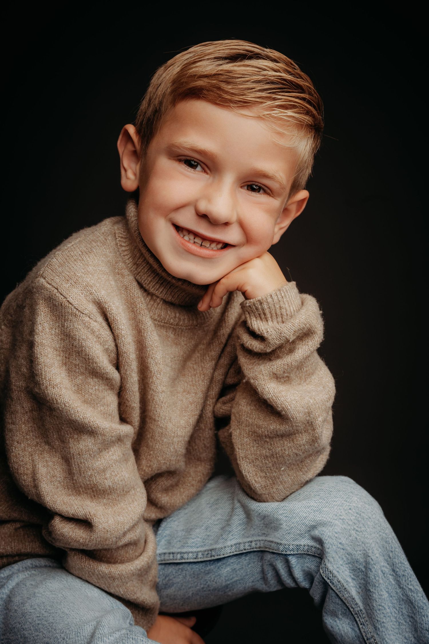 Smiling young boy with blonde hair wearing a beige turtleneck sweater and jeans, posing against a dark background.