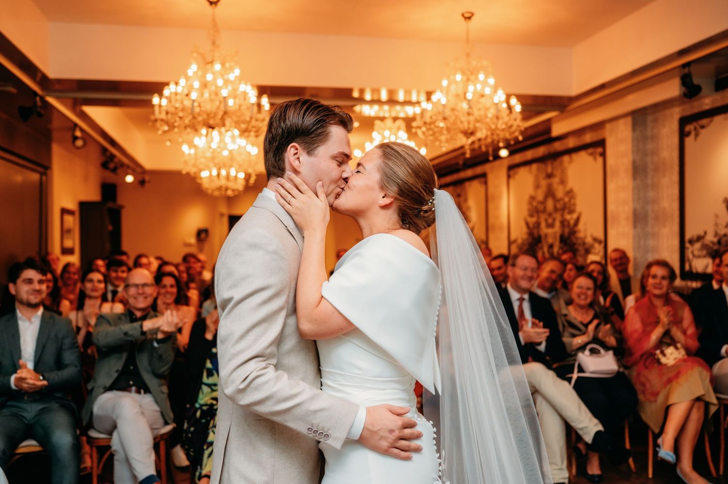 Newlyweds share first kiss at wedding ceremony under golden chandeliers with applauding guests.