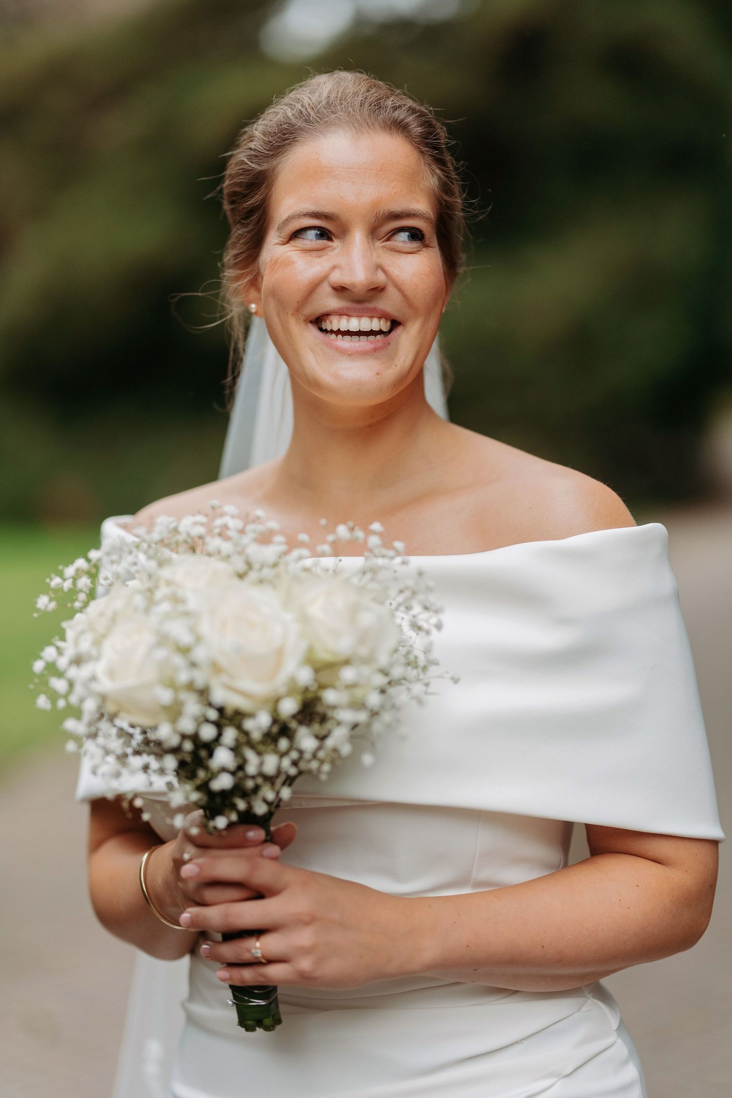 Smiling bride in off-shoulder white gown holds a bouquet of white roses and baby's breath outdoors.