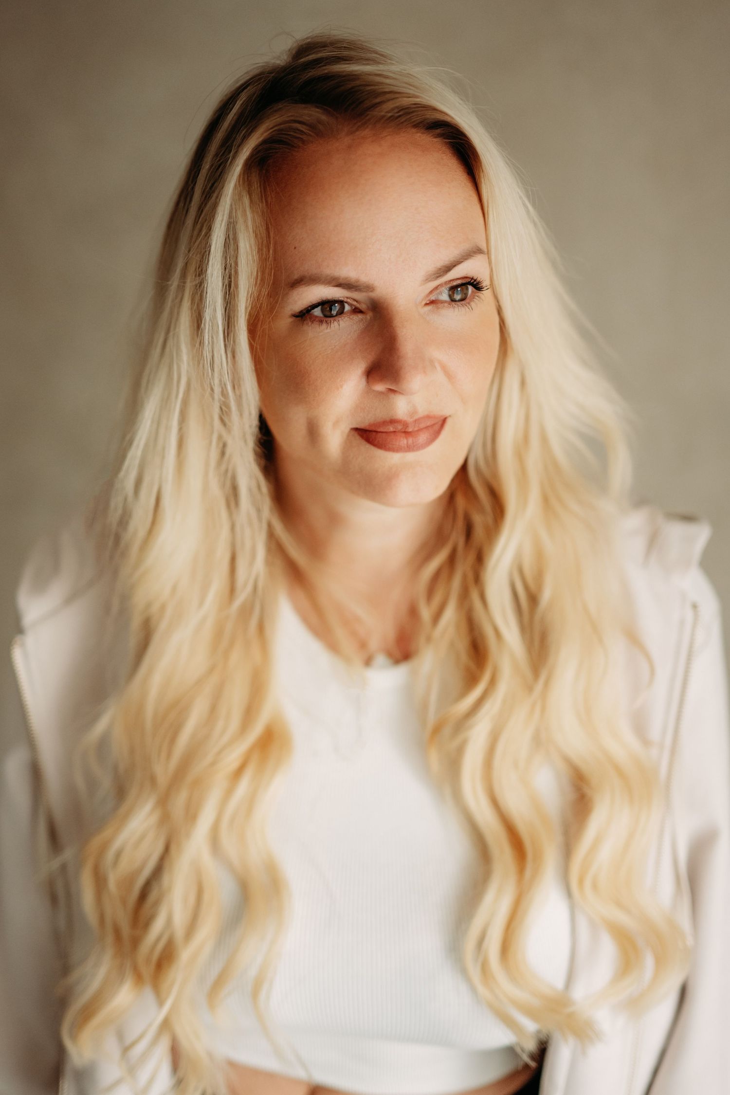 Blonde woman with long wavy hair wearing a white blazer and top, smiling softly against a neutral background.