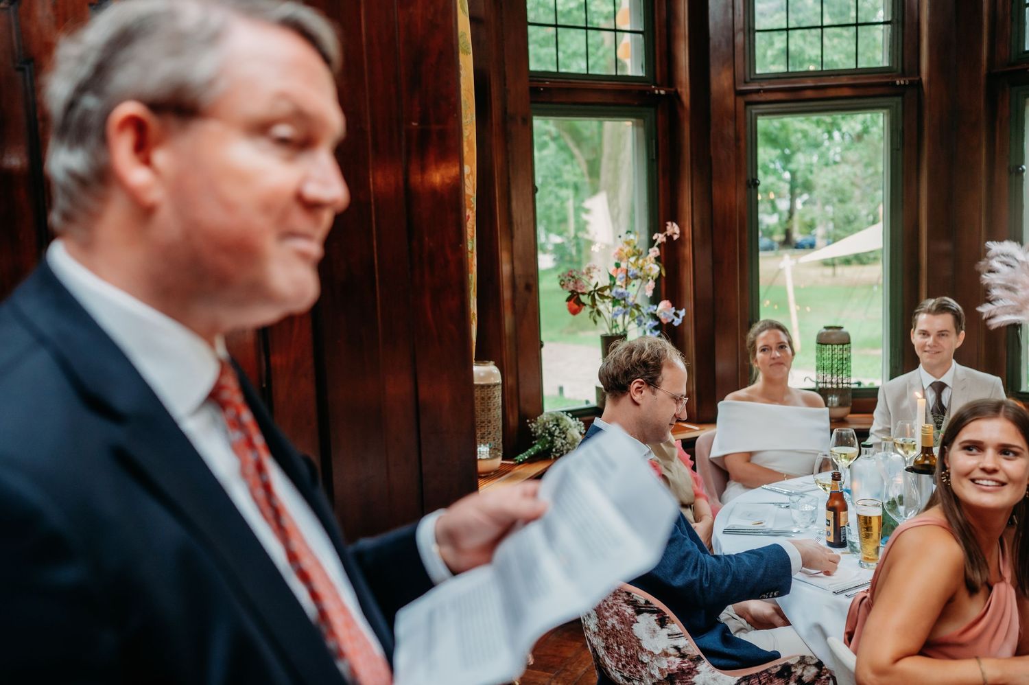 Man giving a speech at an elegant wedding reception in a wood-paneled venue with guests seated at decorated tables.