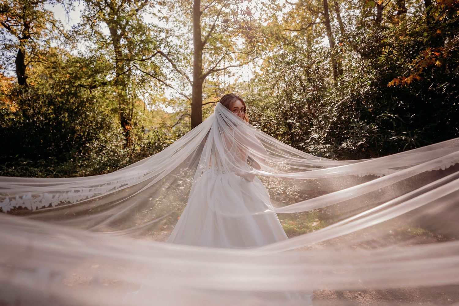 A bride stands in a sunlit autumn forest, her long sheer lace-trimmed veil spread wide across the ground in front of her.