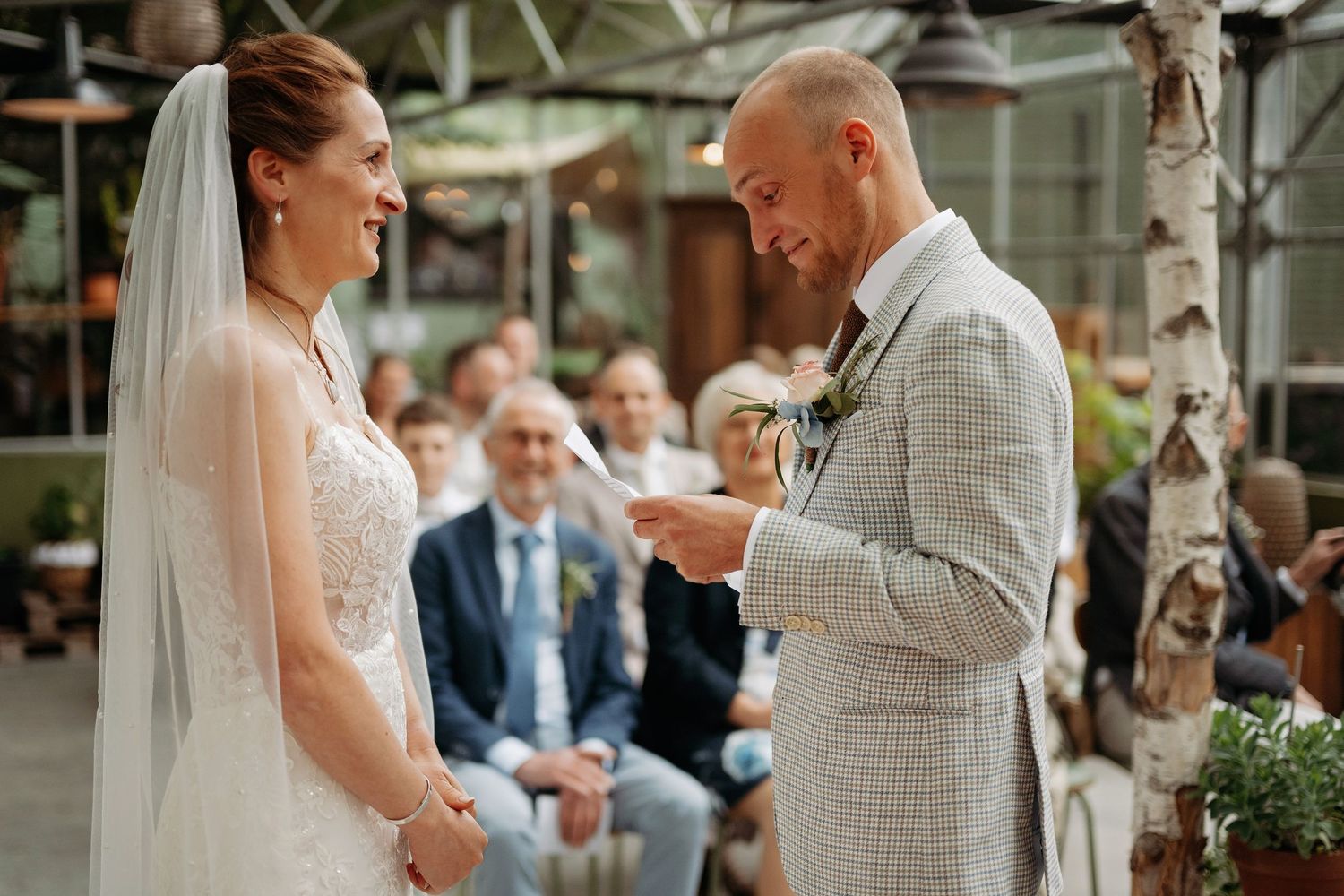 Groom reads wedding vows to smiling bride during indoor ceremony with guests seated behind.
