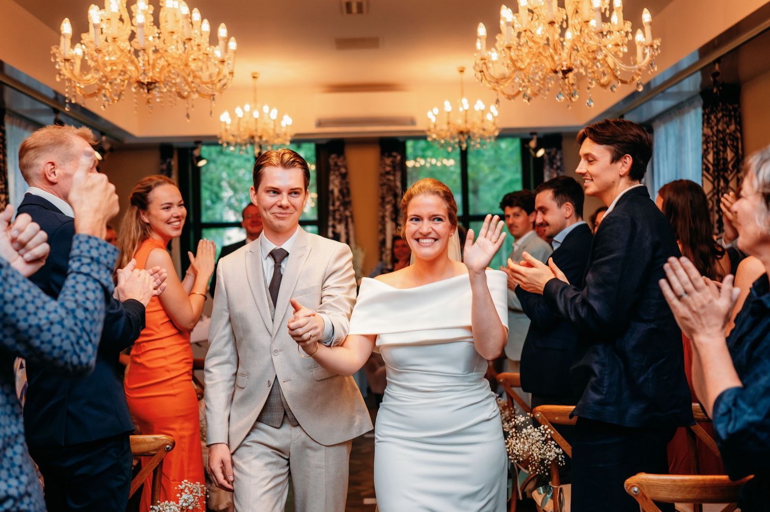 Newlywed couple walks triumphantly through applauding guests in elegant chandelier-lit reception hall.