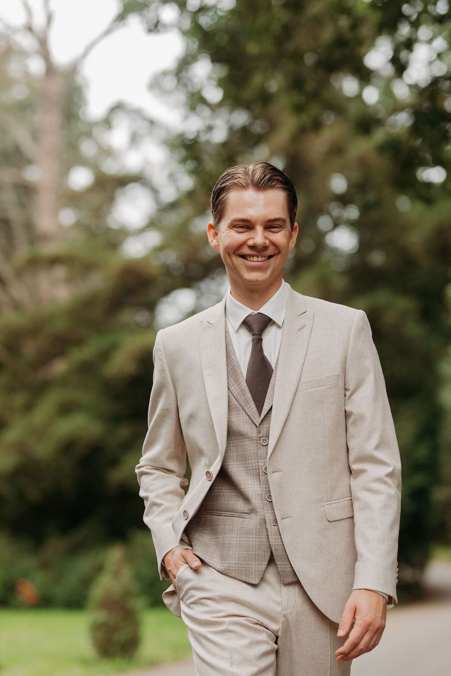 Smiling groom in a beige three-piece suit and gray tie standing outdoors with lush green trees in the background.