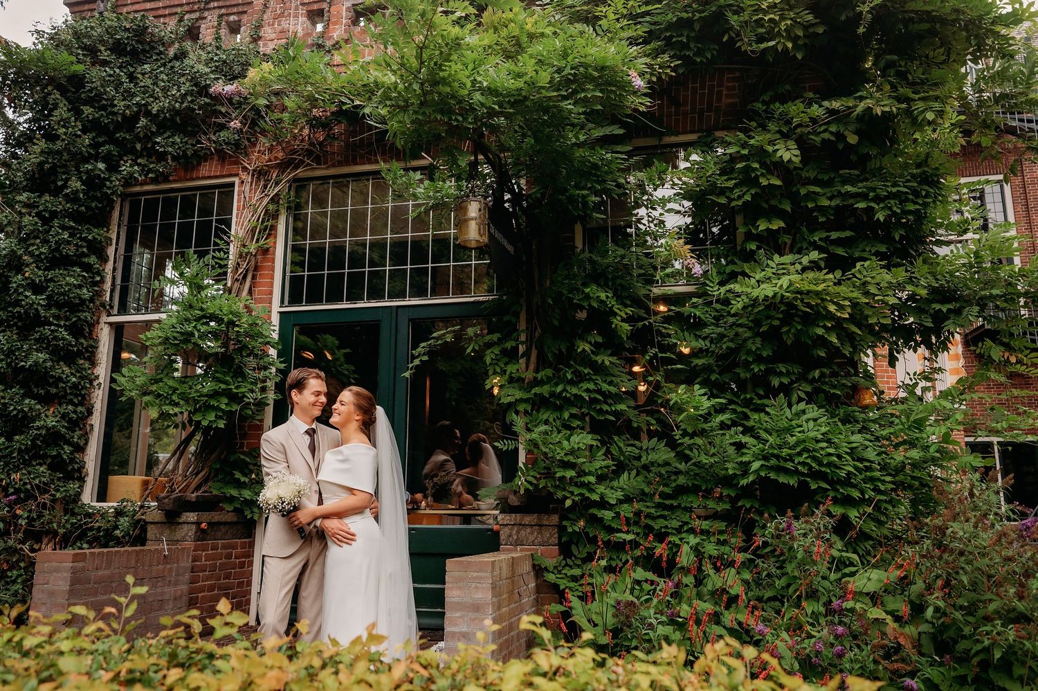 Couple embracing in front of ivy-covered brick building surrounded by lush greenery on their wedding day.