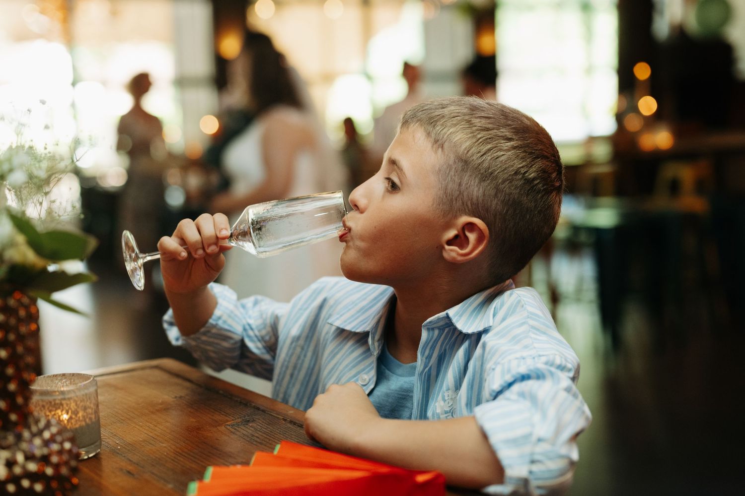 Young boy drinking water from a large wine glass at a restaurant table.