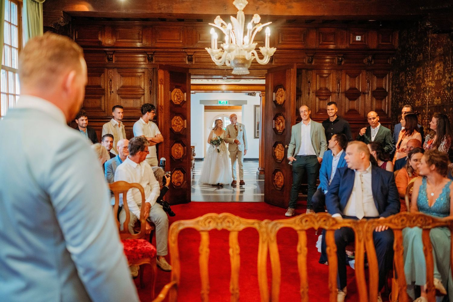 Elegant wedding ceremony in ornate wood-paneled room with chandelier, guests seated as couple stands at doorway.