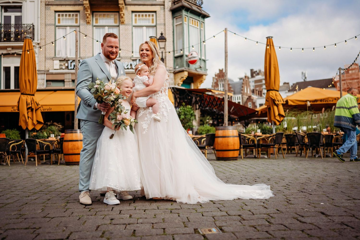 Bride and groom posing with child in European town square, bride in white tulle gown, groom in light blue suit.