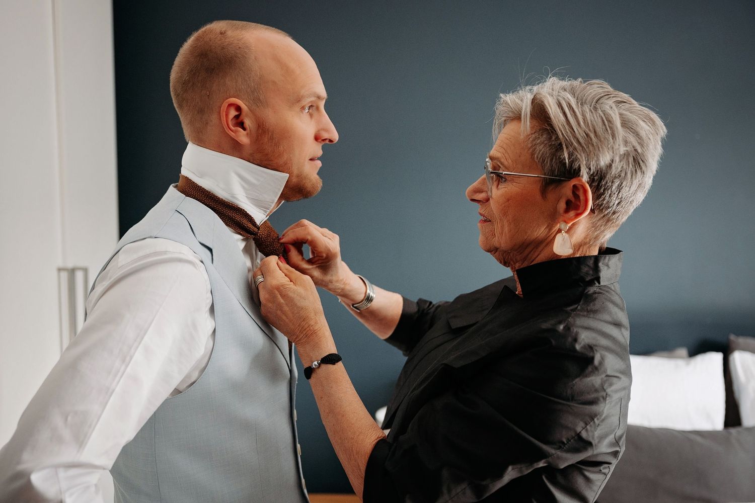 Older woman helping a bald groom tie his brown necktie before the wedding ceremony.