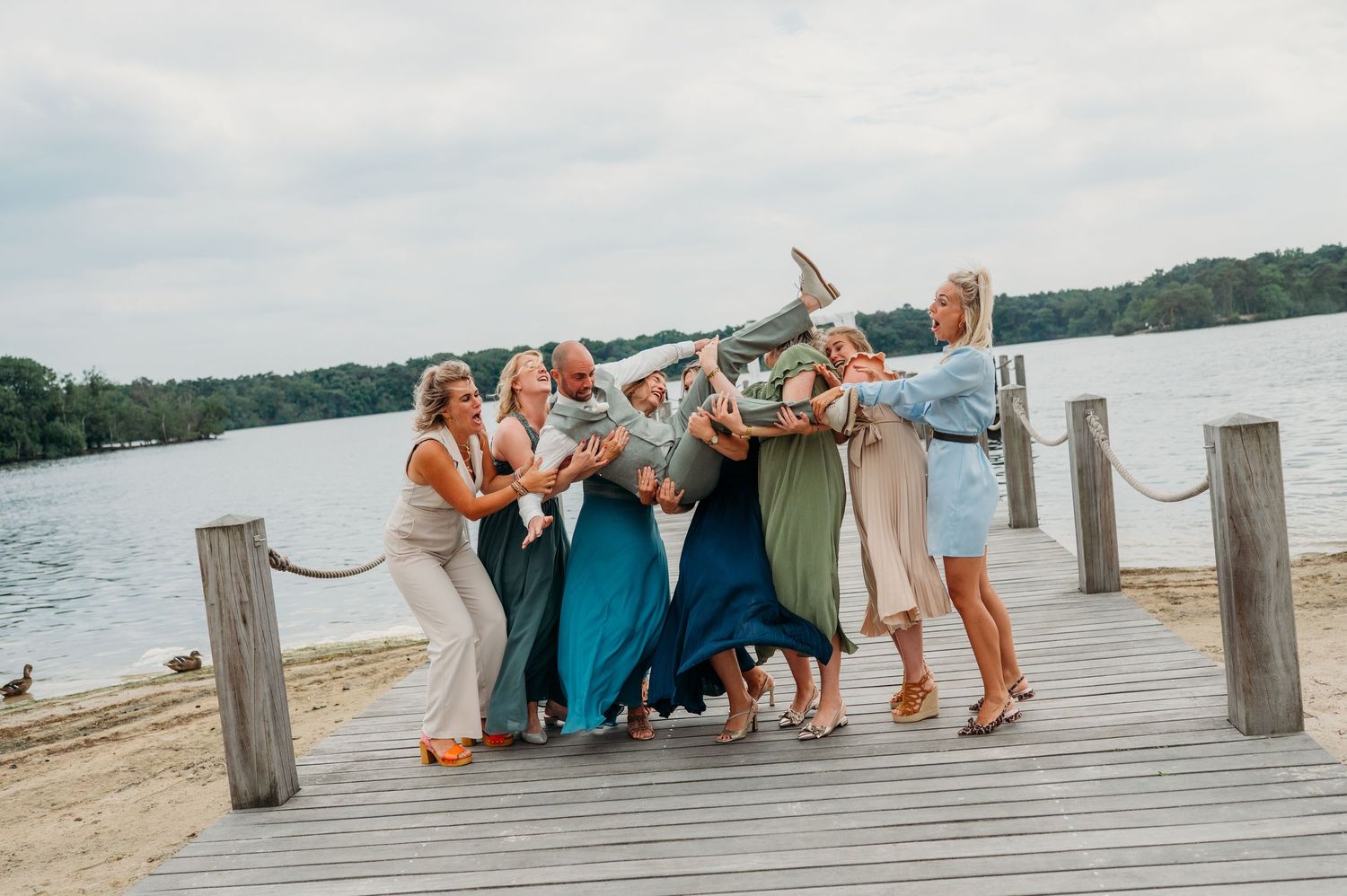 Bruidspaar en gasten poseren op houten steiger bij meer, bruidsfotografie IJM IJzereman Vught, Brabant.