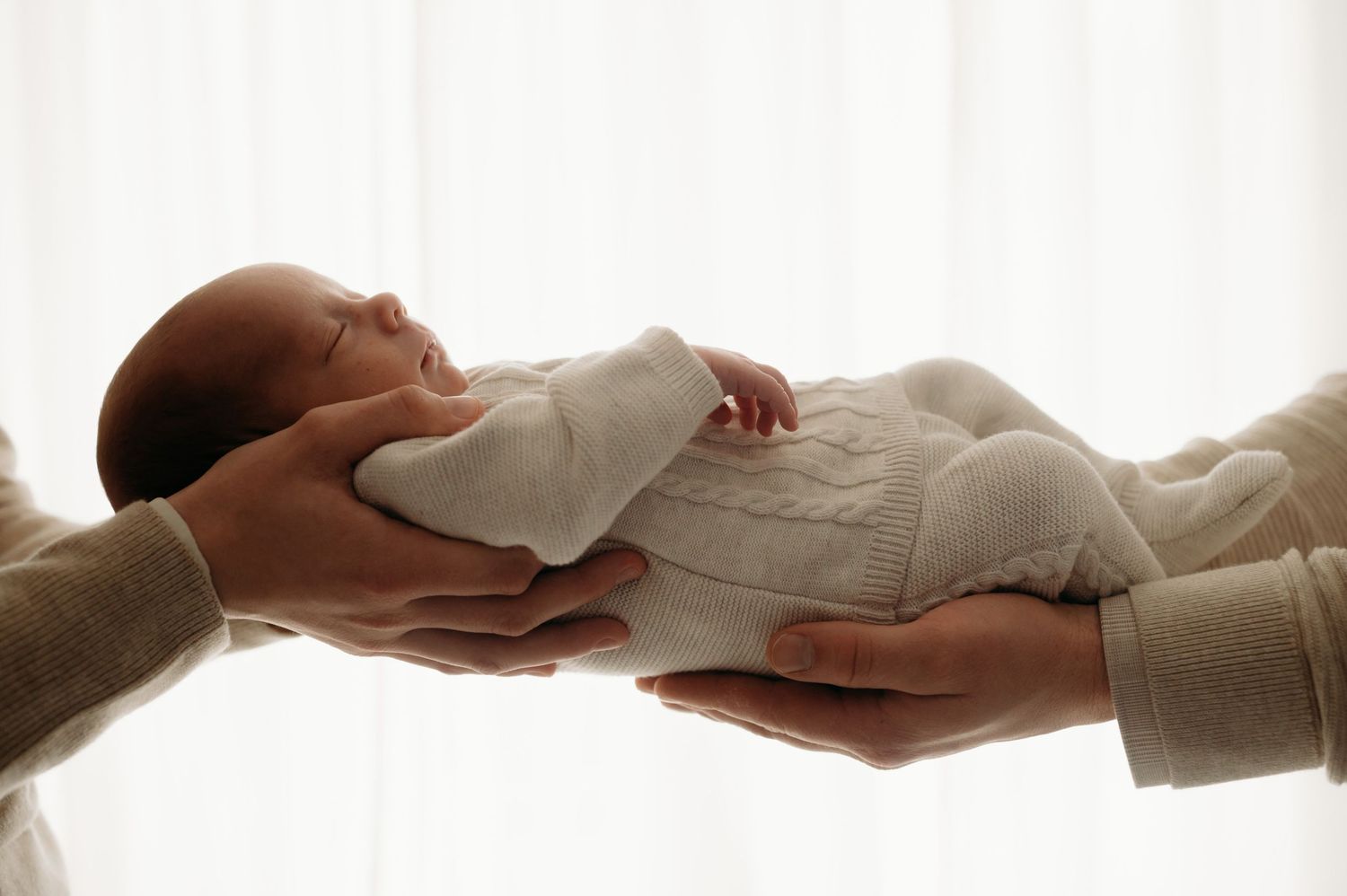 Zachte handen houden pasgeboren baby in witte kleding tegen helder raamlicht, newbornfotografie Brabant