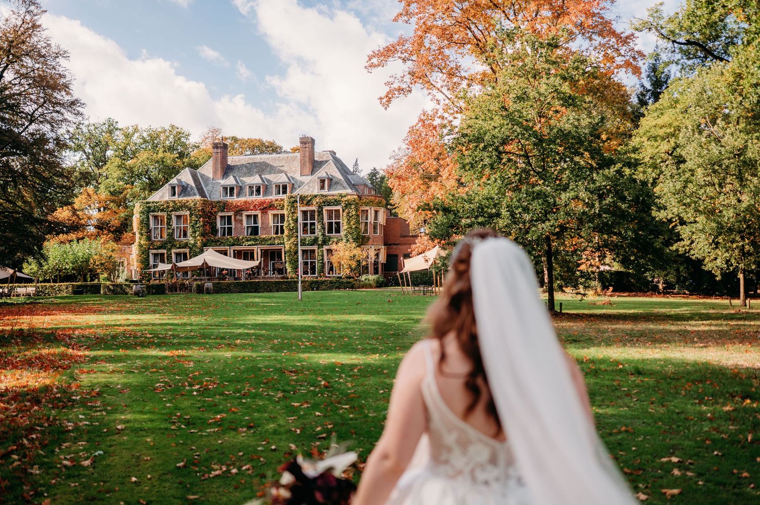 Bride in a white lace gown and long veil faces a historic manor house across a sunlit lawn framed by colorful autumn trees.