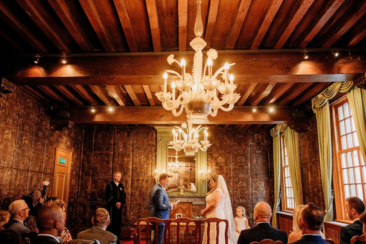 Elegant wedding ceremony in a historic room with wooden ceiling, chandelier, and couple exchanging vows before guests.