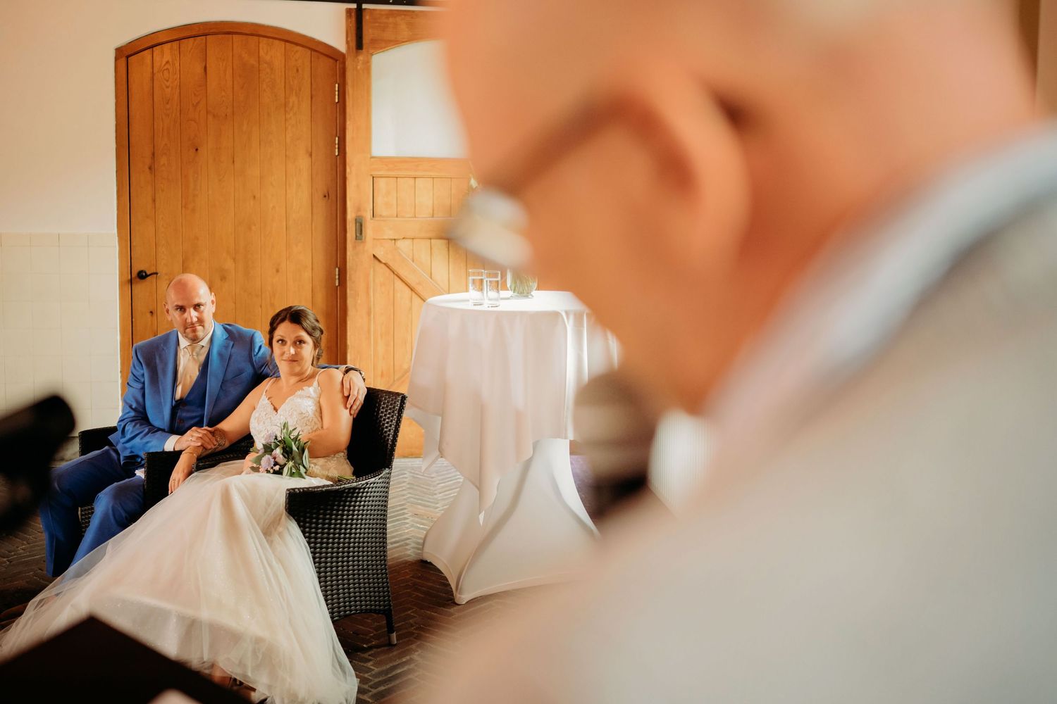 Bride and groom in blue suit seated at wedding ceremony listening to officiant at altar.