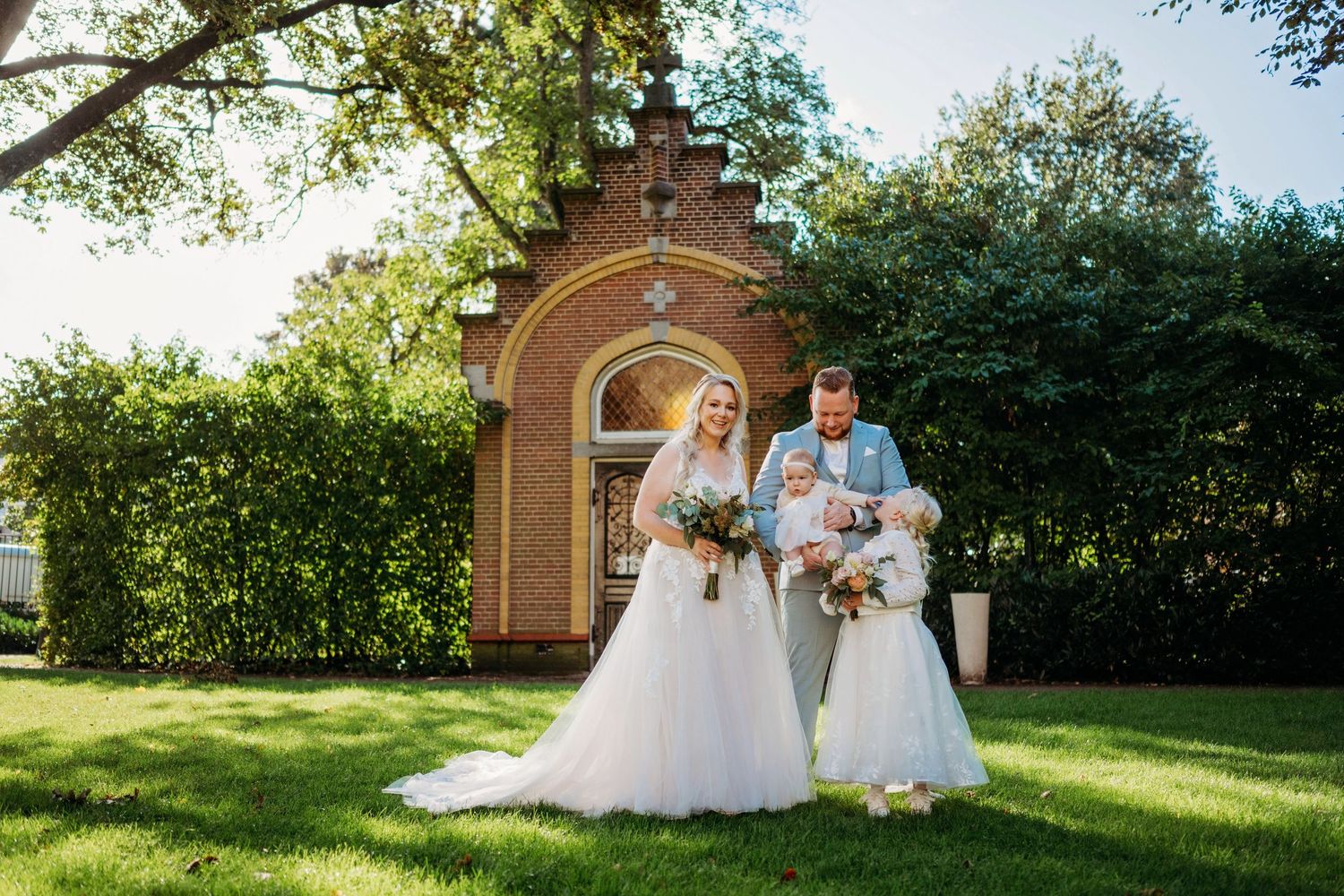Bride and groom with children pose on green lawn before brick chapel on sunny wedding day.
