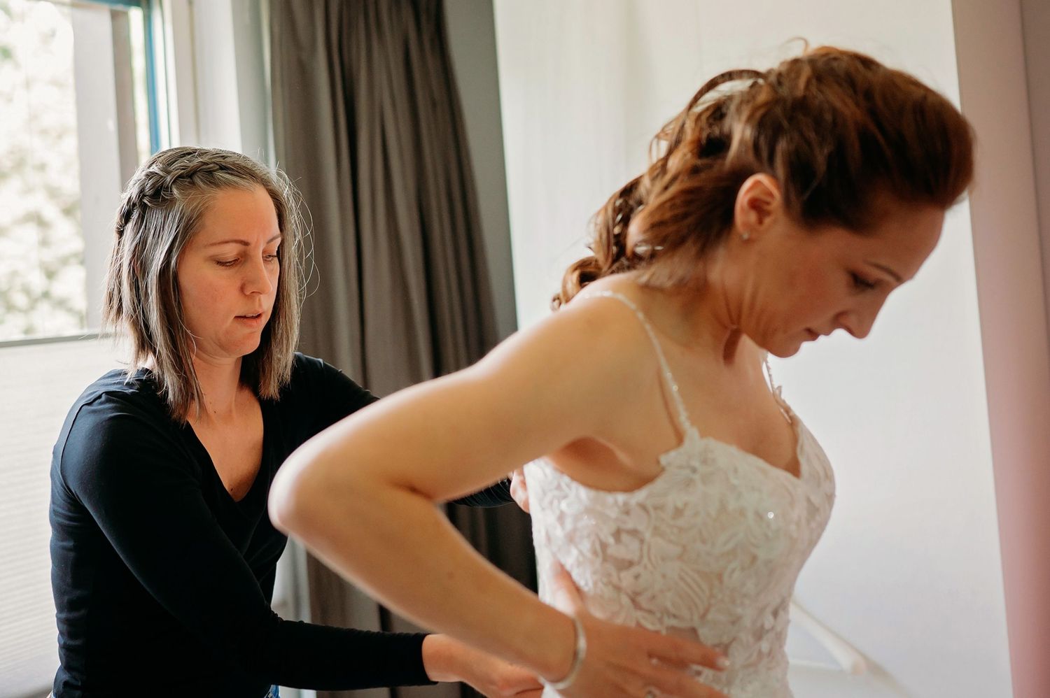 A bridesmaid helps a bride in a white lace wedding gown get dressed, adjusting the back of her gown indoors.