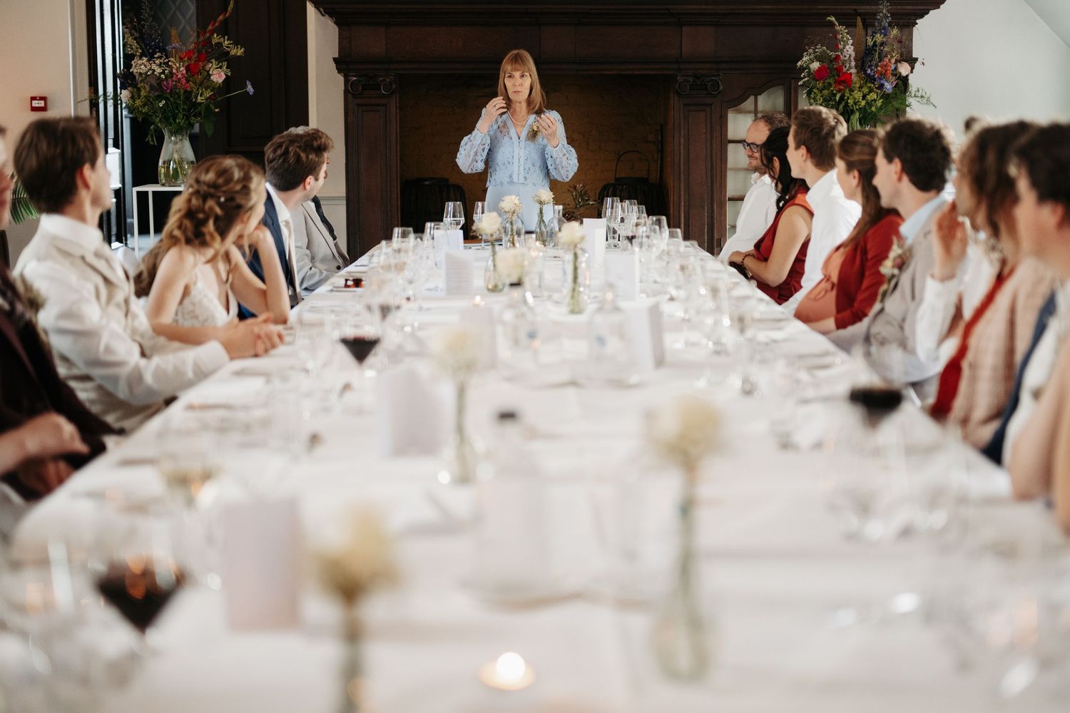 A woman gives a speech at an elegant long dining table surrounded by guests at a formal wedding reception.