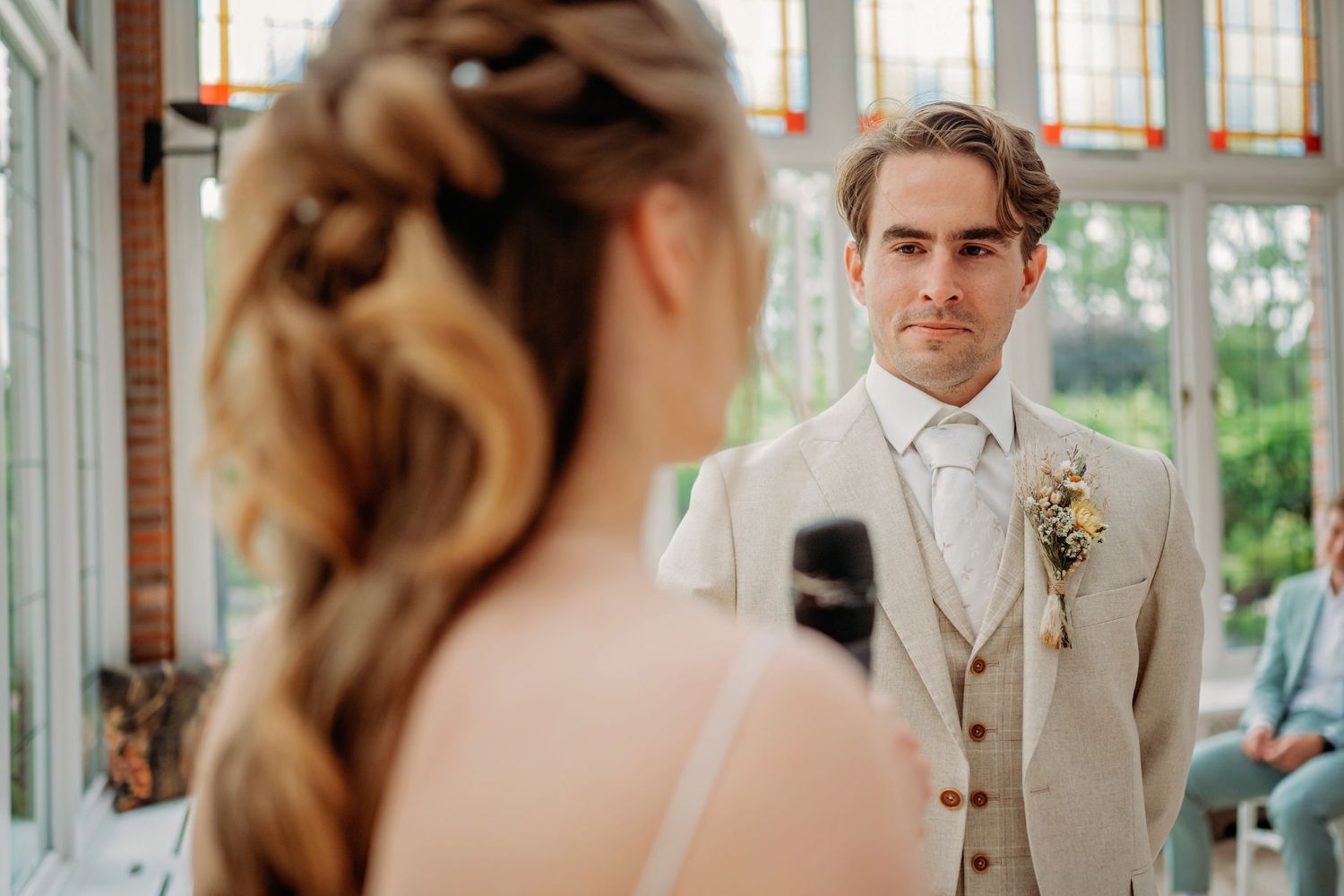 Groom in beige suit gazes at bride during intimate indoor wedding ceremony with floral boutonniere.