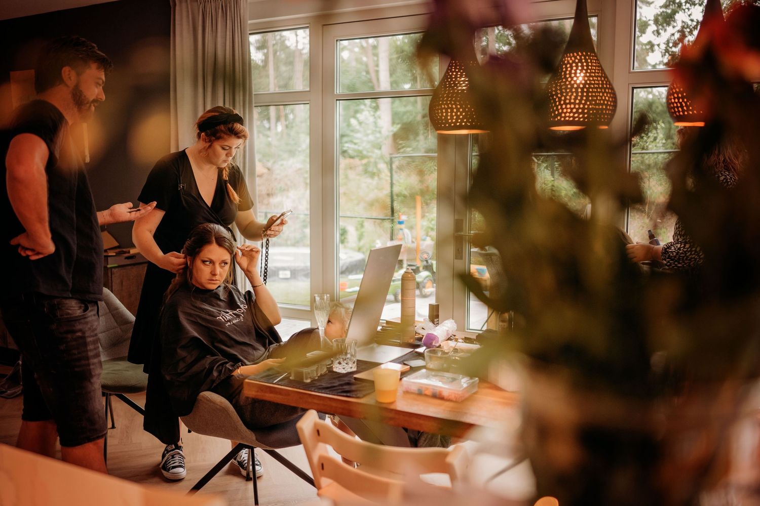 Hairstylist preparing bride's hair in a warmly lit room with pendant lights and a laptop on a wooden table.