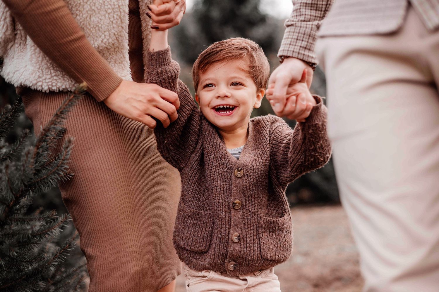 A joyful toddler in a brown cardigan laughs while being held by both hands by parents outdoors near evergreen trees.
