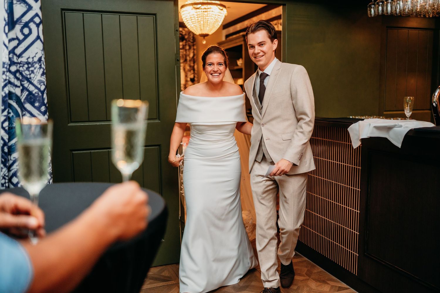 Smiling bride in white off-shoulder gown and groom in tan suit entering wedding reception as guest holds champagne flute.