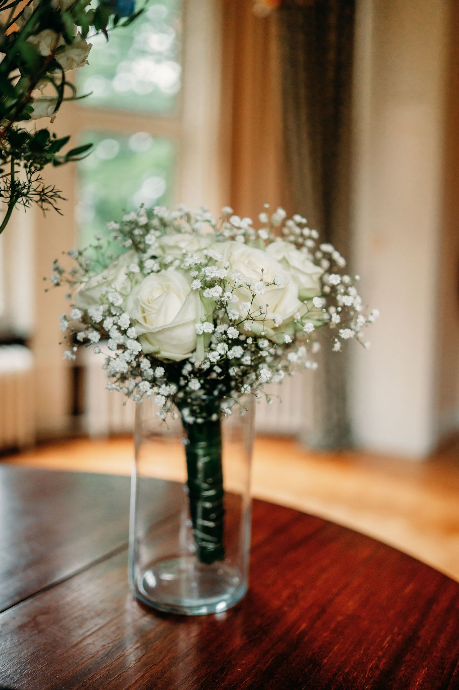 Elegant bridal bouquet of white roses and baby's breath in a glass vase on a wooden table near a window.