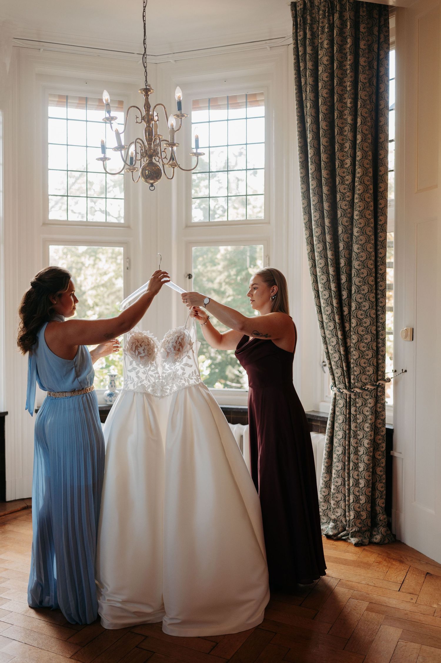 Two bridesmaids in blue and burgundy dresses hold up a white lace wedding gown in a bright room with a chandelier and tall windows.