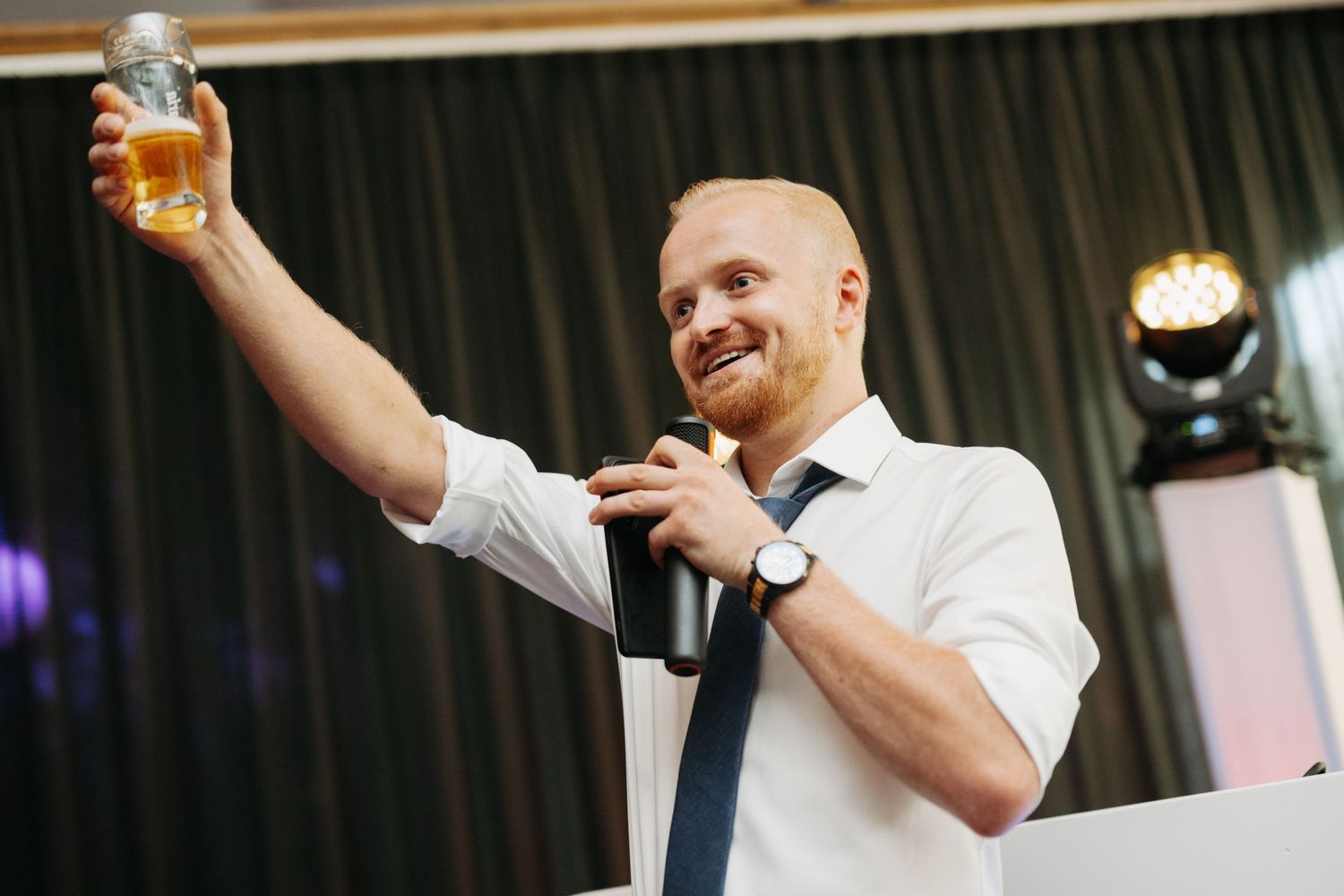 Smiling man in white shirt and tie raising a glass beer mug while holding a microphone at an event.