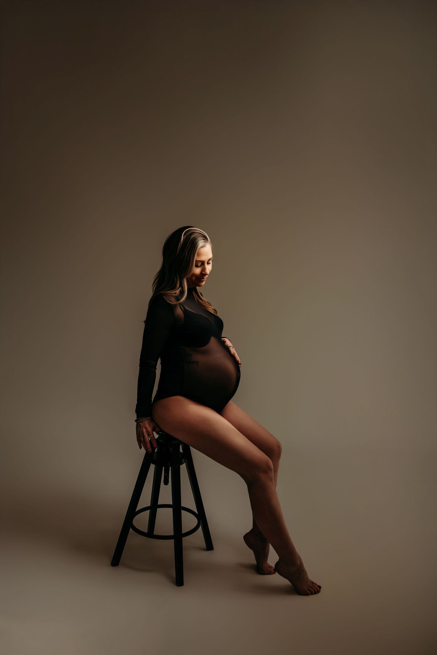 Artistieke boudoirfoto van zwangere vrouw zittend op houten kruk met donkere achtergrond, zwangerschapsfotografie Brabant.