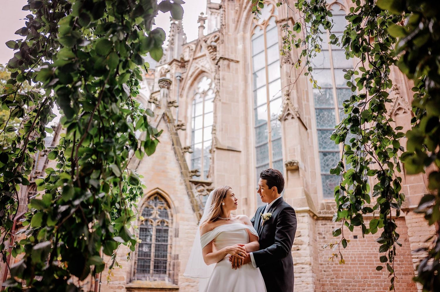 Bruidspaar omarmt elkaar voor gotische kerk met klimop en sierlijke architectuur, bruidsfotografie Noordbrabants Museum Den Bosch, Brabant.