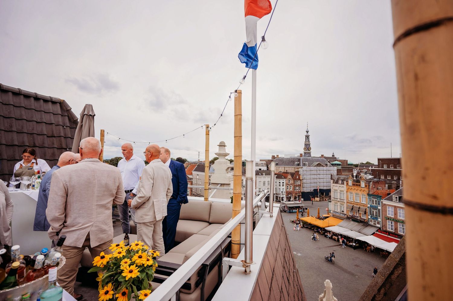 Guests mingle on a rooftop terrace overlooking a European city square, with a Dutch flag and yellow flowers nearby.