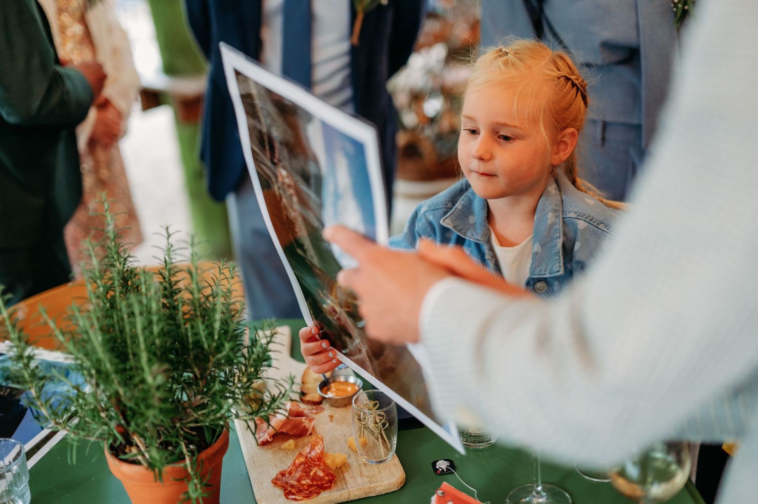 A young girl with braided hair looks at a photo print at an outdoor event table with potted herbs and food.