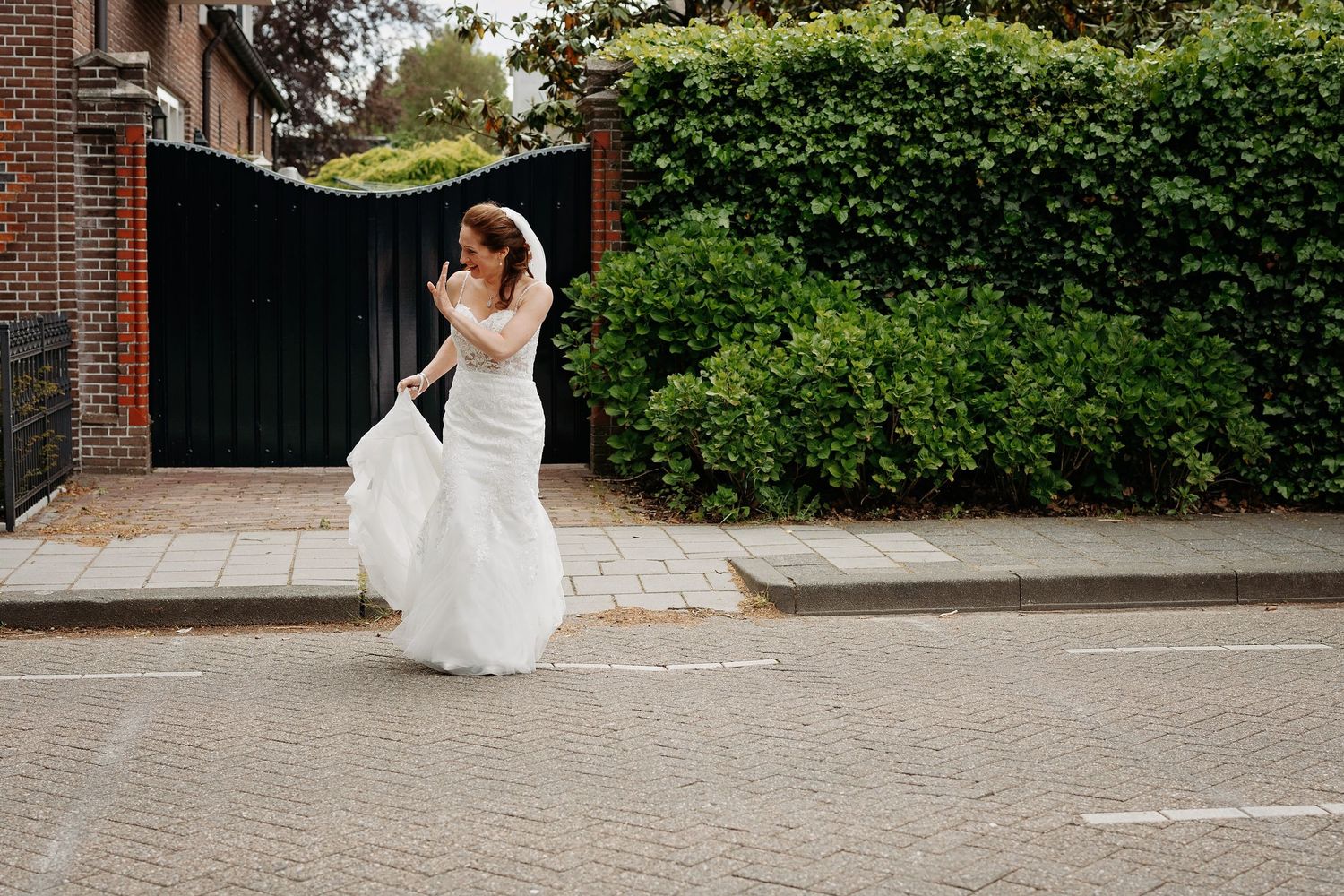 Bride in white wedding gown holding veil on a cobblestone street beside lush green hedges and a black gate.
