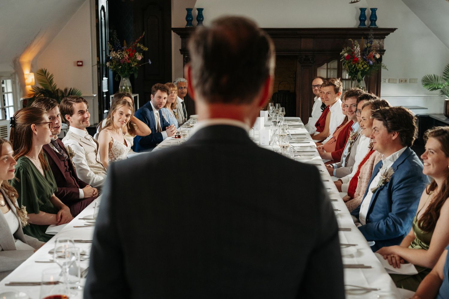 A man in a suit addresses guests seated at a long elegant dining table during a formal wedding reception.