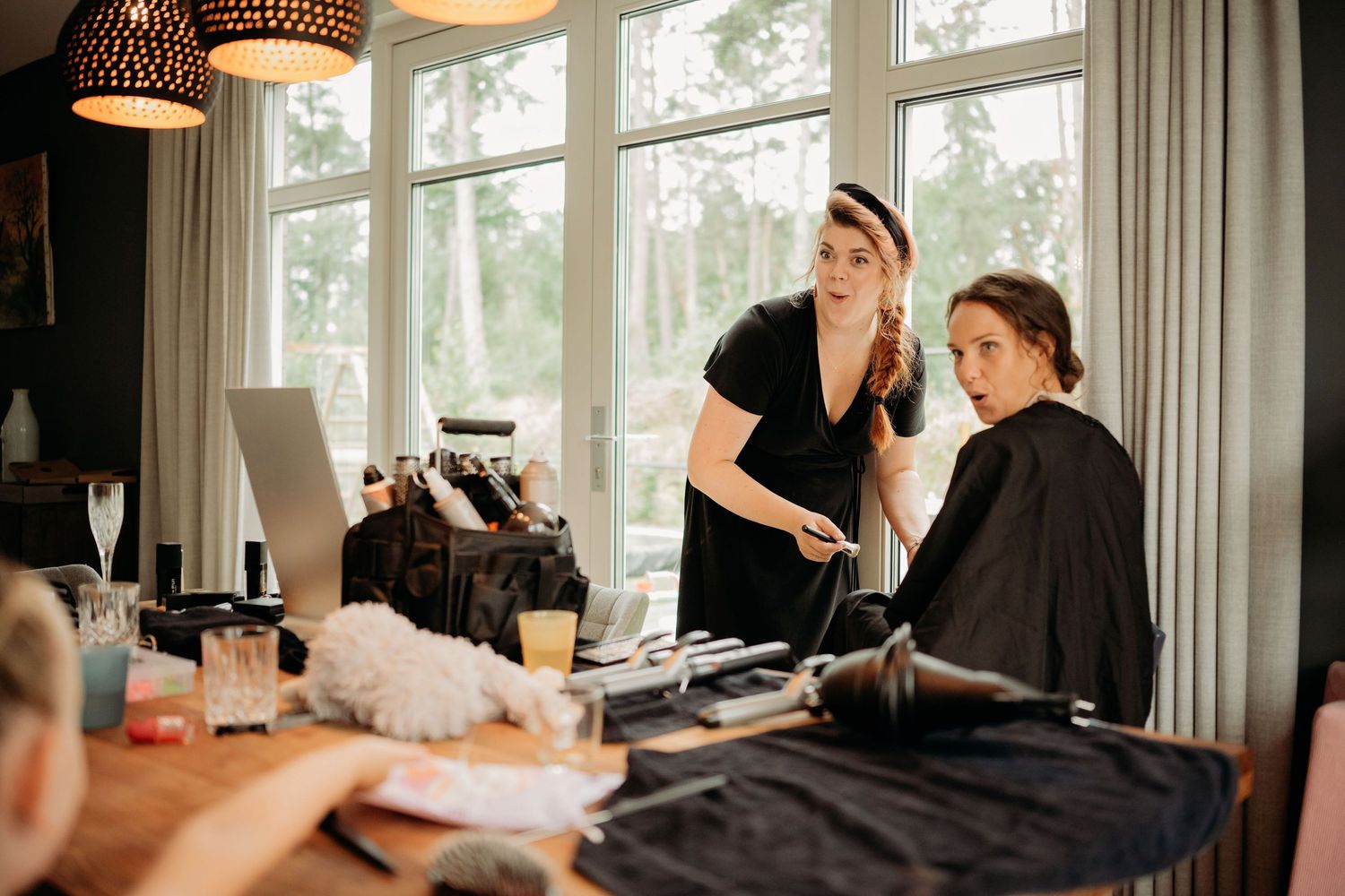 Hair stylist styling a seated woman's hair in a bright room with large windows and styling tools on a table.