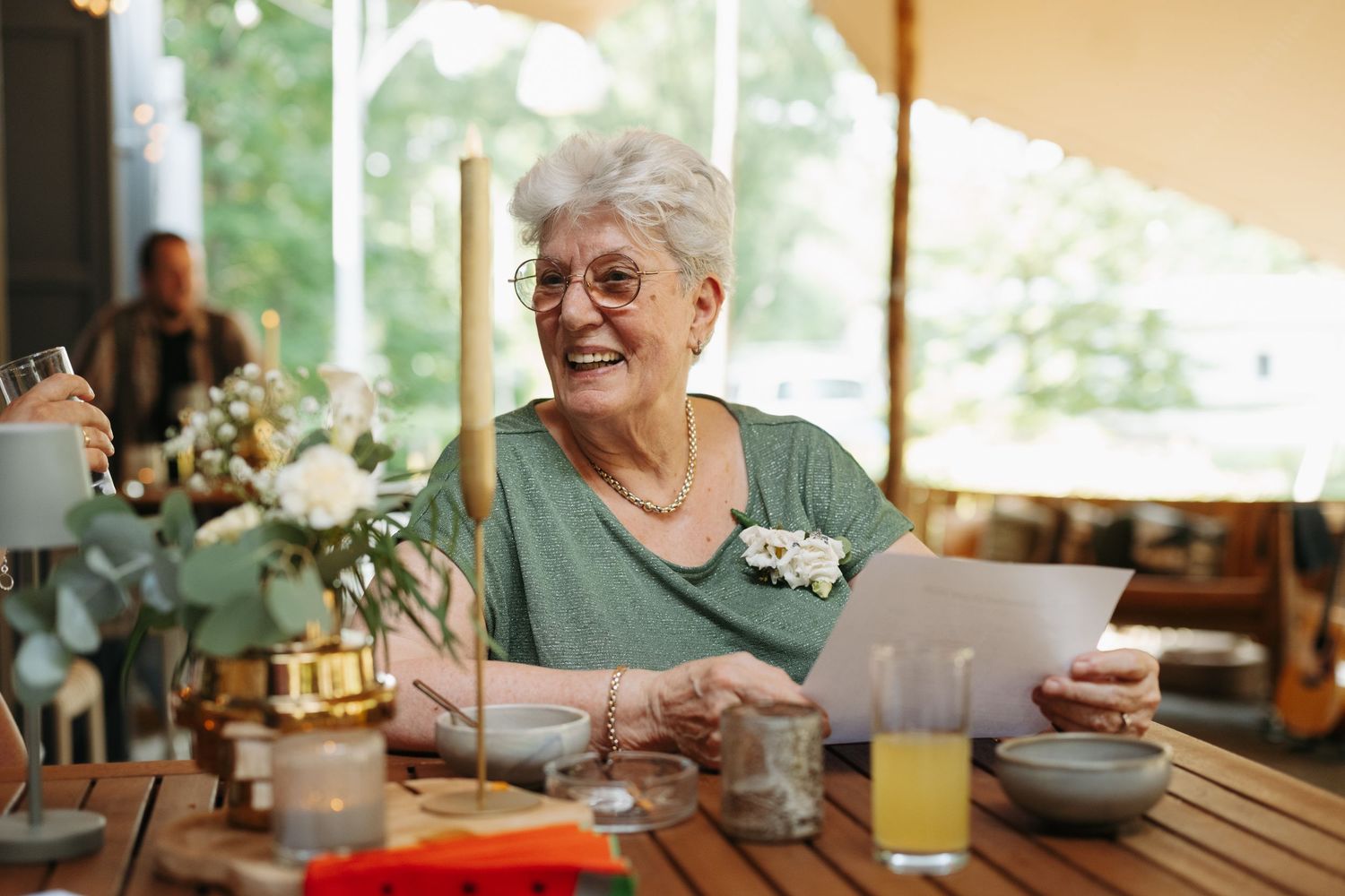 Happy elderly woman smiling while reading a paper at an outdoor wedding reception.