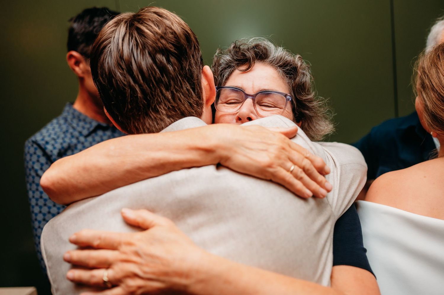 Two people sharing an emotional hug at a gathering, with a man in a blue shirt visible in the background.