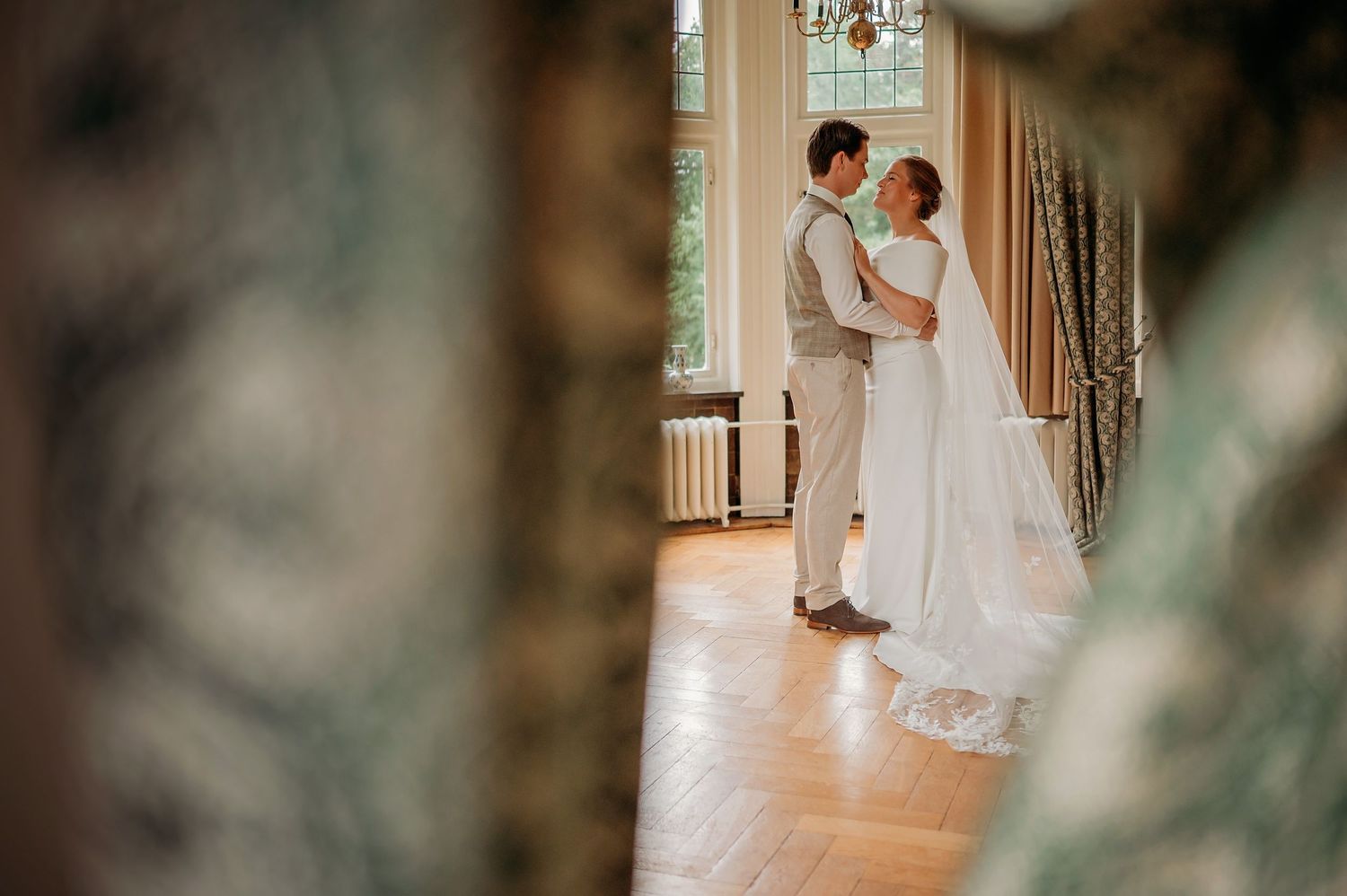 Bride and groom share intimate moment in elegant sunlit room with hardwood floors.
